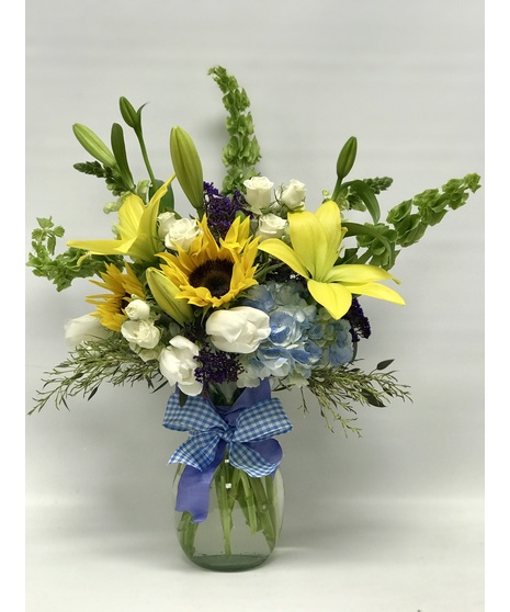 Yellow and blue flowers in a clear glass vase accompanied by a stuffed bear and a mylar balloon that says 