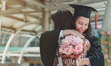 A graduate in a cap and gown embraces another person holding a bouquet of pink and white flowers, within an outdoor setting featuring a structure with beams.